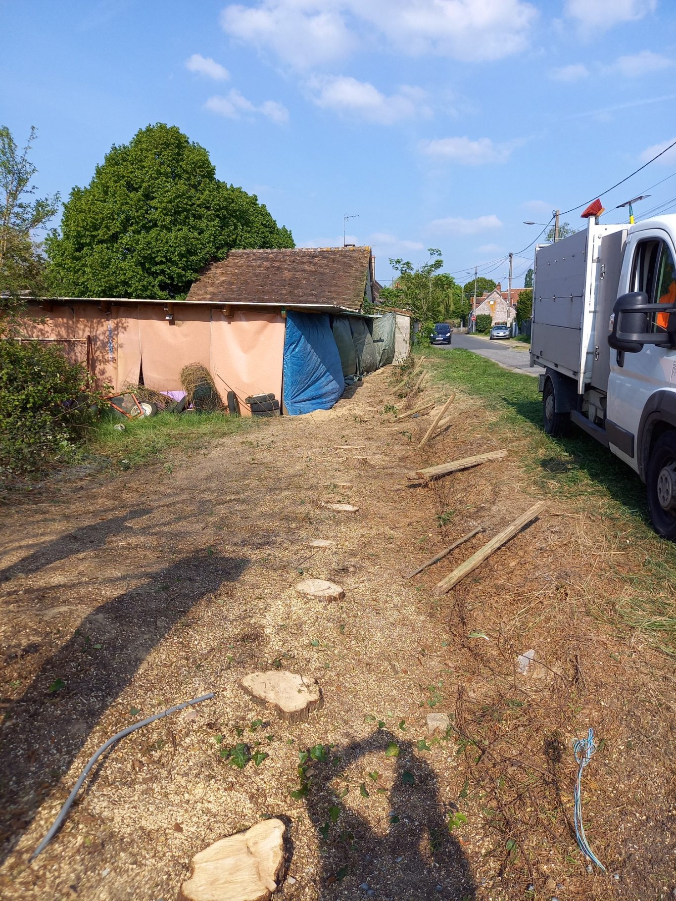 Abattage d'une haie Vue d'un chemin de terre avec une maison en arrière-plan et un véhicule stationné.