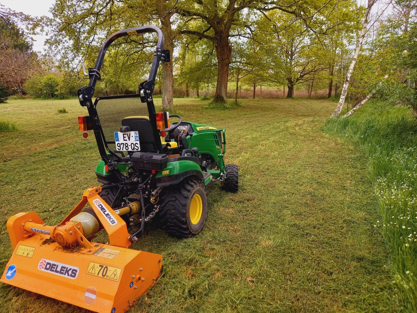 Fauche de hautes herbes Tracteur équipé d'une tondeuse, sur un terrain gazonné avec des arbres en arrière-plan.