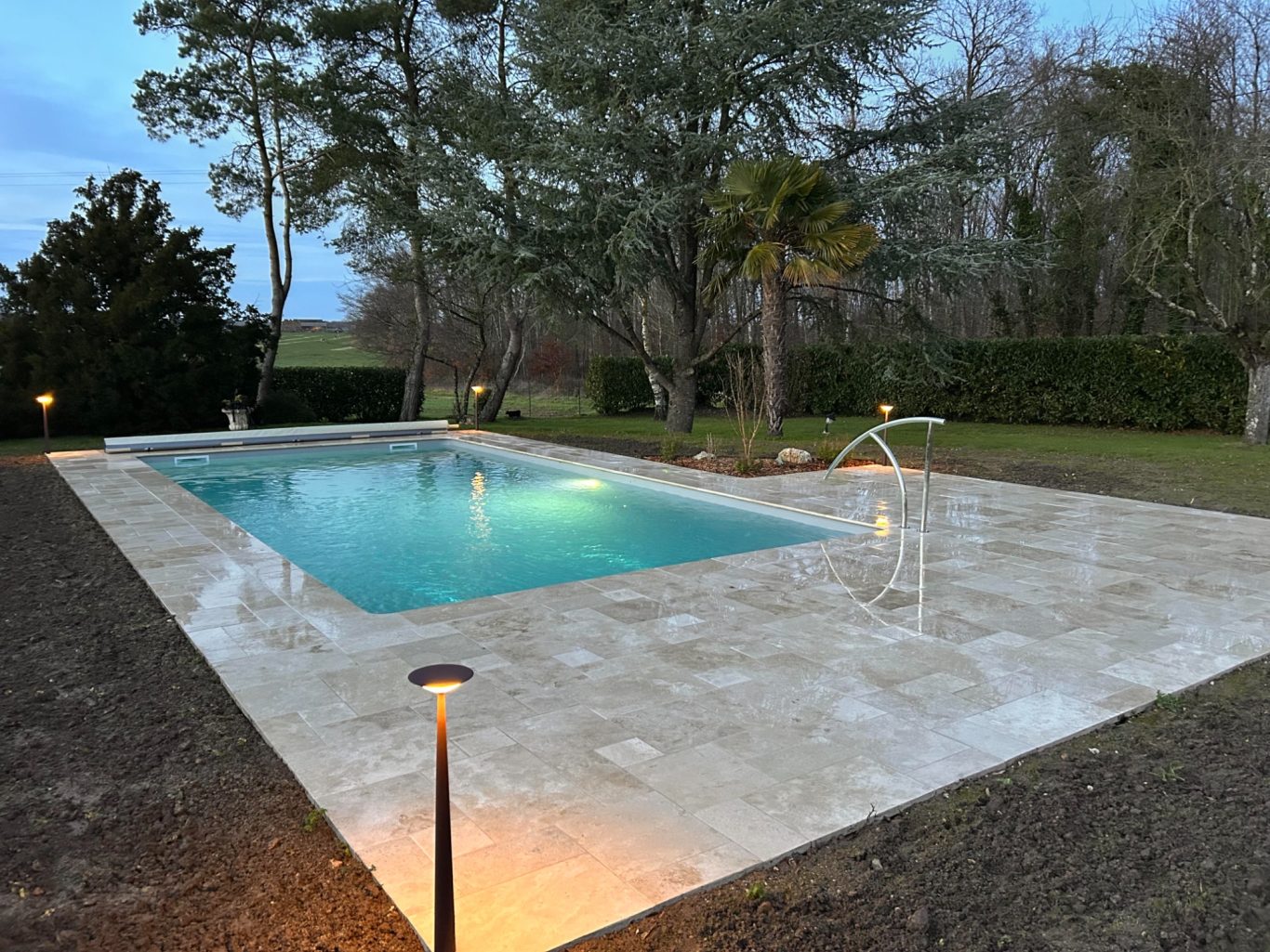 Eclairage / Rambarde Piscine entourée d'une terrasse en pierre, éclairée, avec des arbres en arrière-plan.