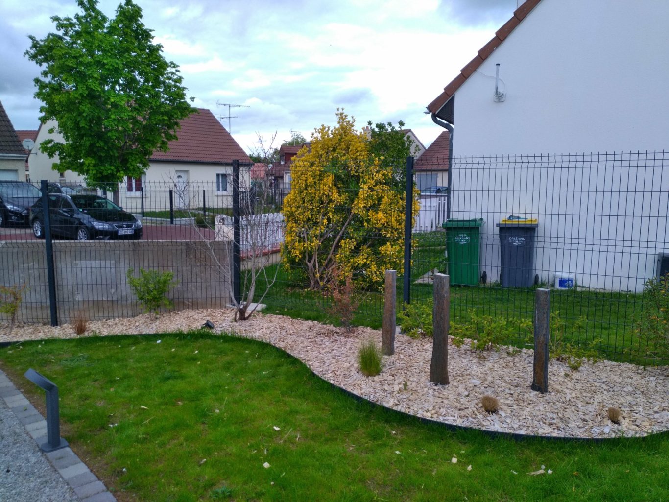 Massif avec palis de schiste Jardin avec gravier, plantes et trois poteaux en bois, sur fond de maisons.