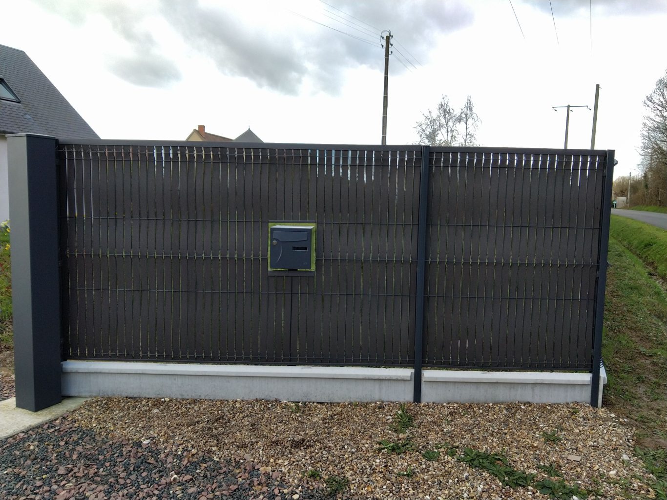 Boite aux lettres encastrée Clôture en métal noir avec un panneau de contrôle sur le côté. Piste en gravier devant.