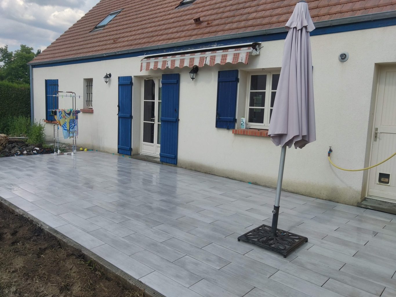 Terrasse en carrelage Terrasse en pavés gris devant une maison avec parasol et volets bleus.