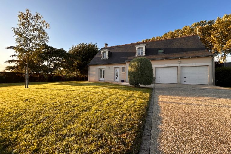 Maison en pierre avec garage, jardin verdoyant et ciel bleu en arrière-plan.