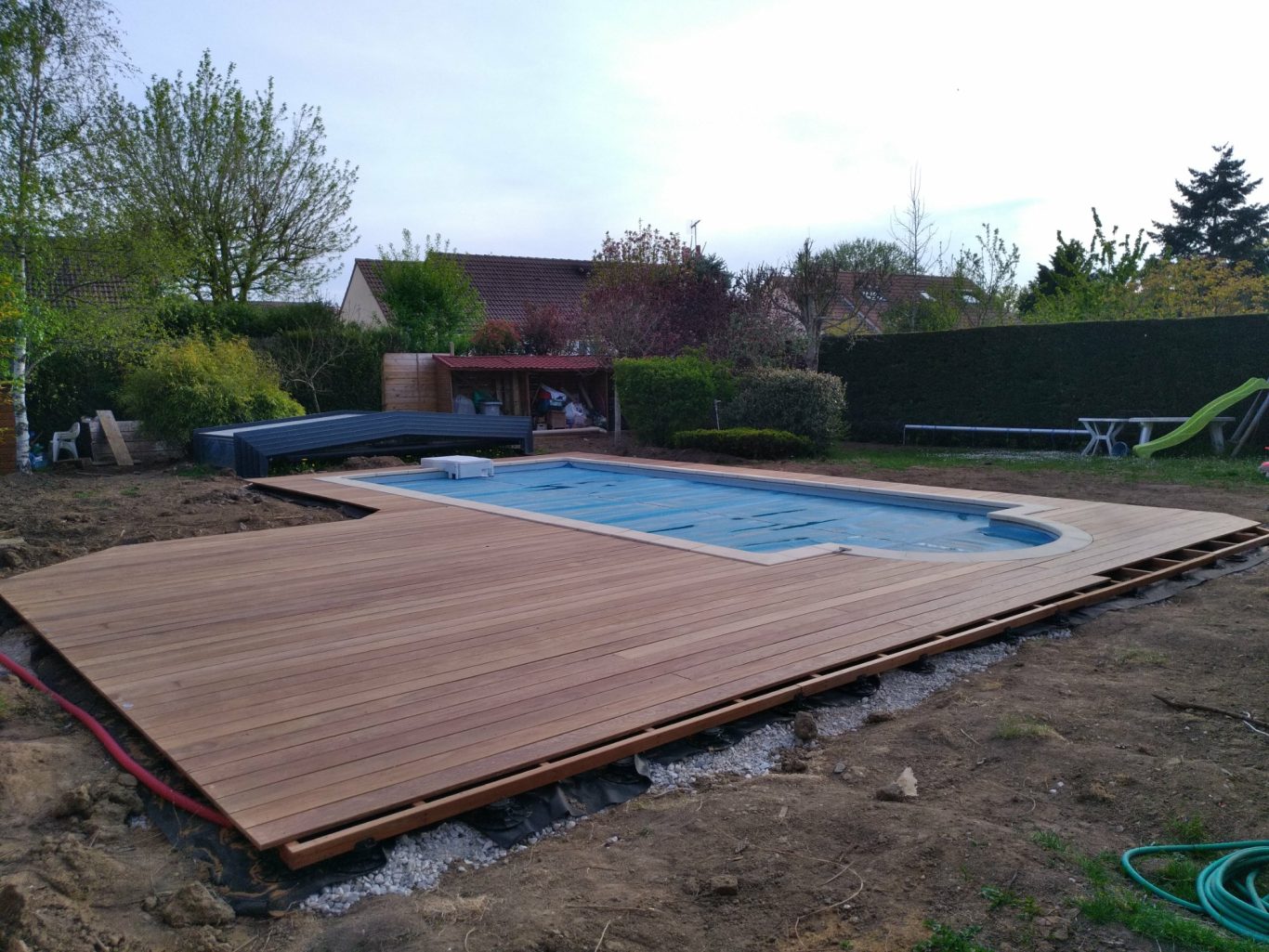 Préparation des lames de rives Une piscine entourée d'une terrasse en bois, avec un jardin verdoyant en arrière-plan.