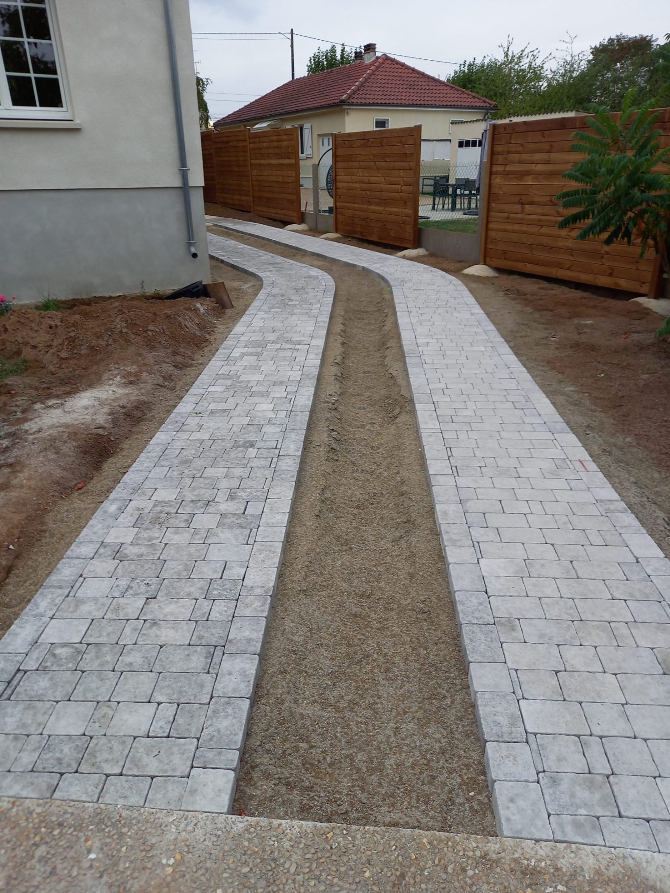 Avant de l'allée pavée Allée en pavés avec une section en gravier, bordée de clôtures en bois.