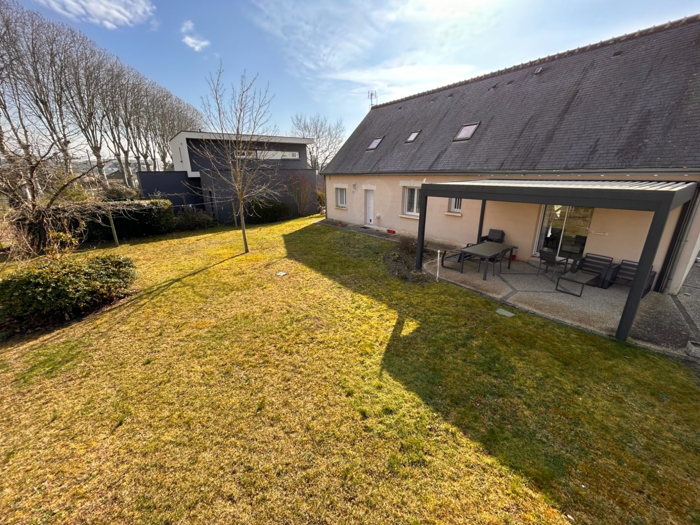 Début de chantier Maison avec jardin ensoleillé et terrasse, entourée d'arbres. Ciel bleu en arrière-plan.