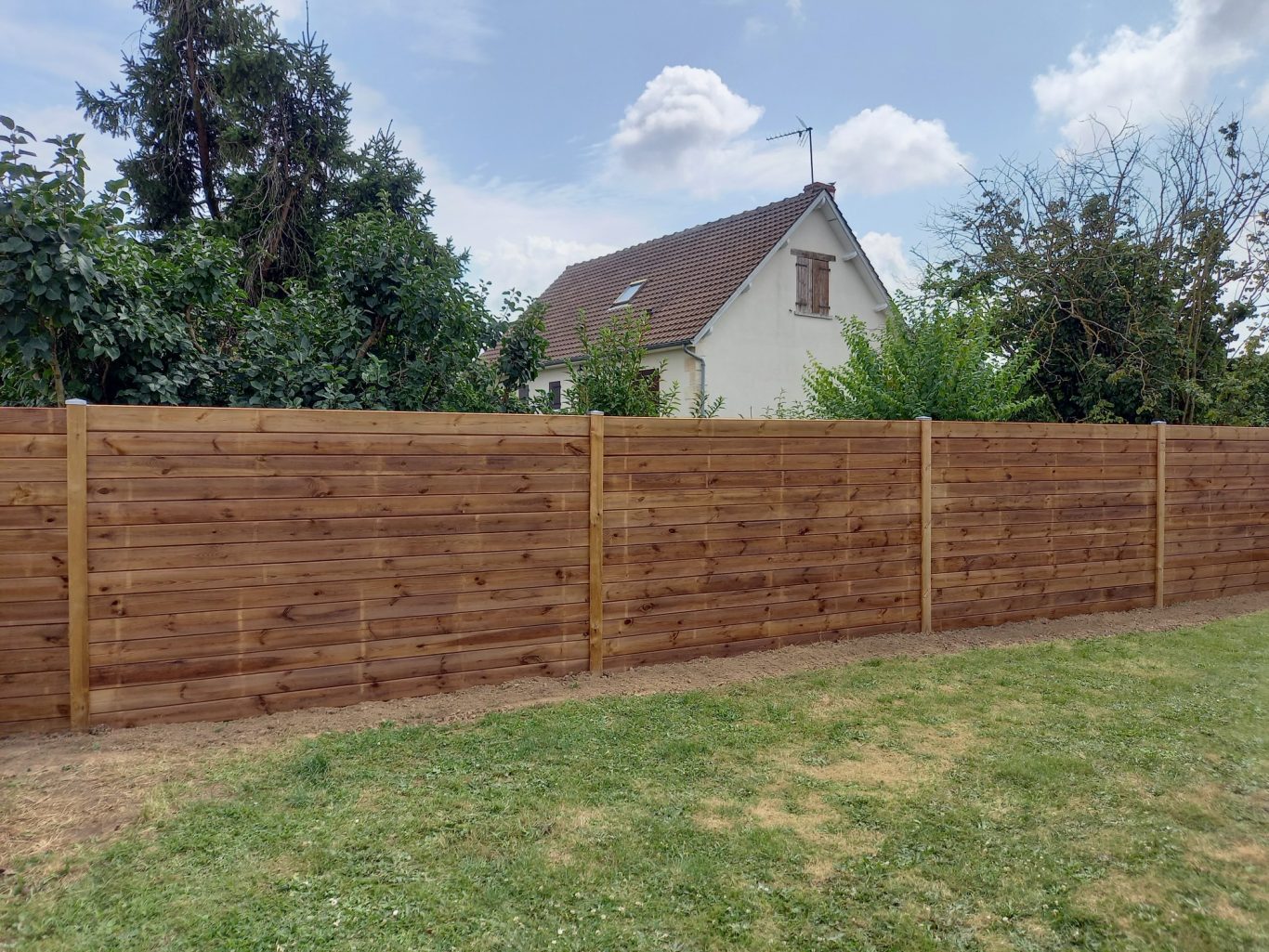 Clôture en panneaux bois Clôture en bois bordant un jardin avec une maison à l'arrière-plan. Ciel dégagé.