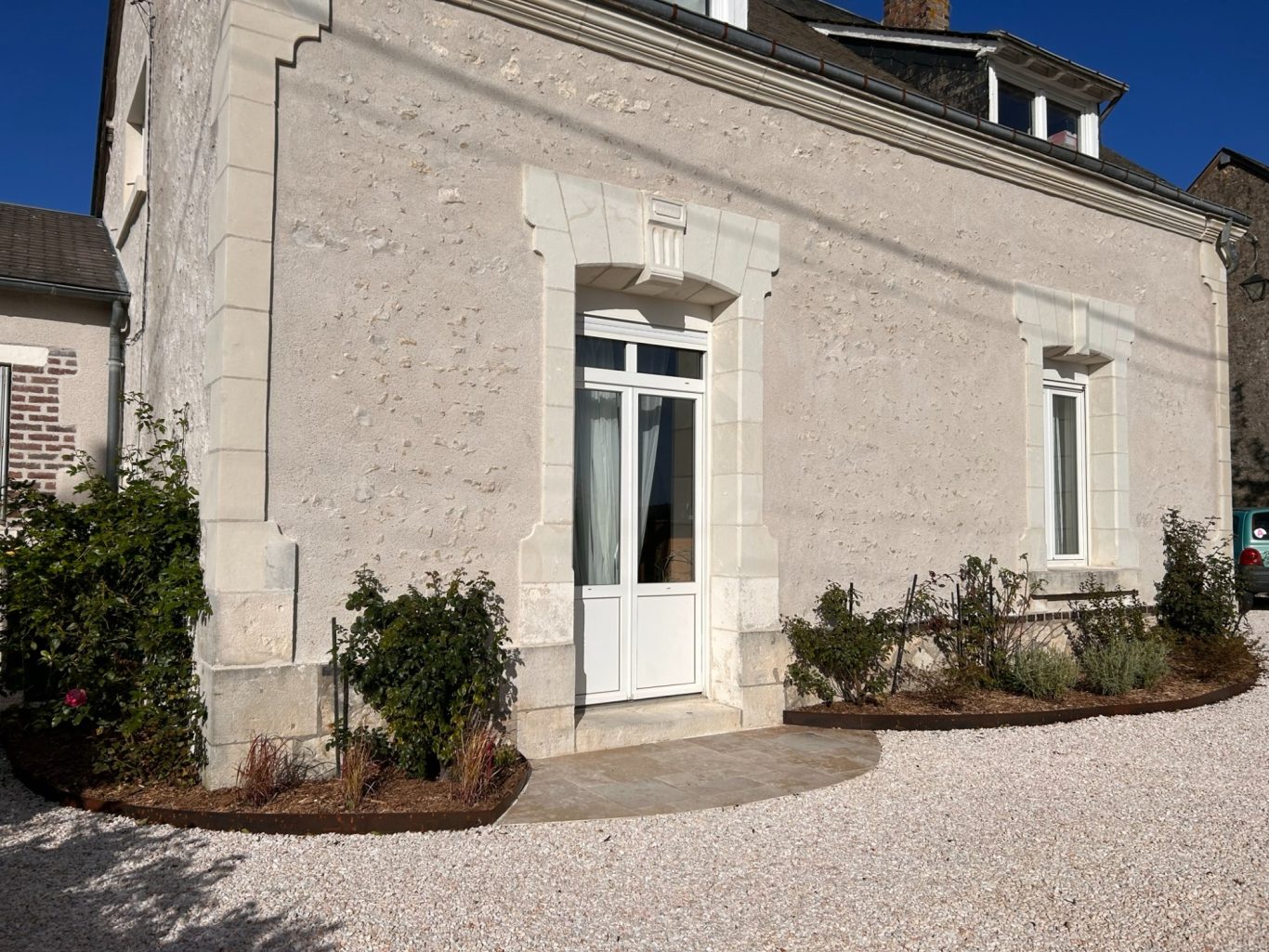 Cours / Massif / Pas de porte en travertin Bâtiment en pierre avec une entrée blanche, jardin soigné et gravier à l'avant.