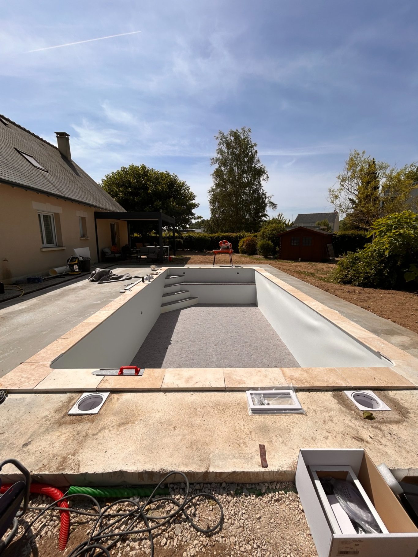 Pose des margelles / Préparation liner Piscine en cours de construction avec terrasse et jardin environnant. Ciel dégagé.