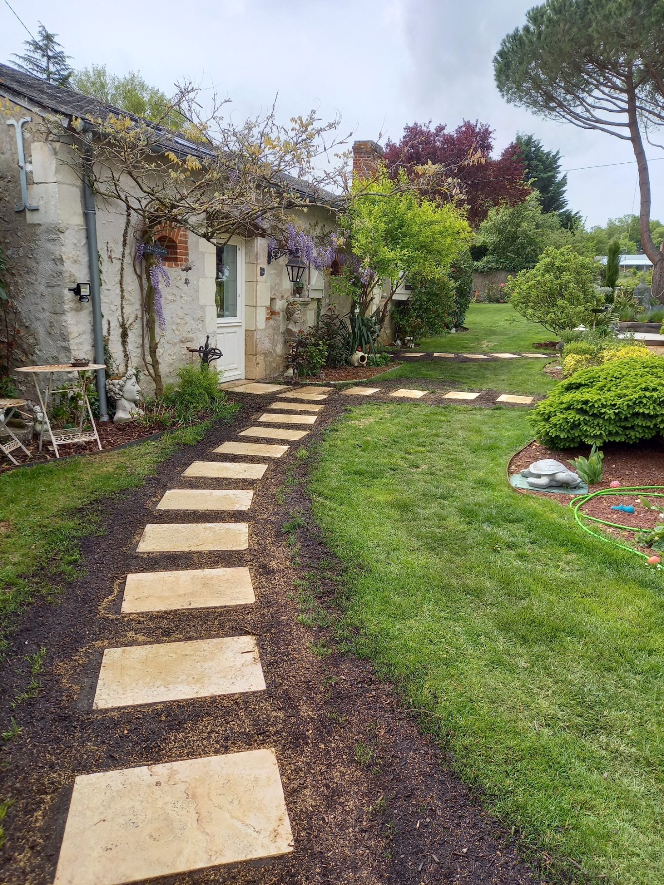 Pas japonais en travertin Chemin en dalles de pierre menant à une maison, entouré de verdure et de plantes.