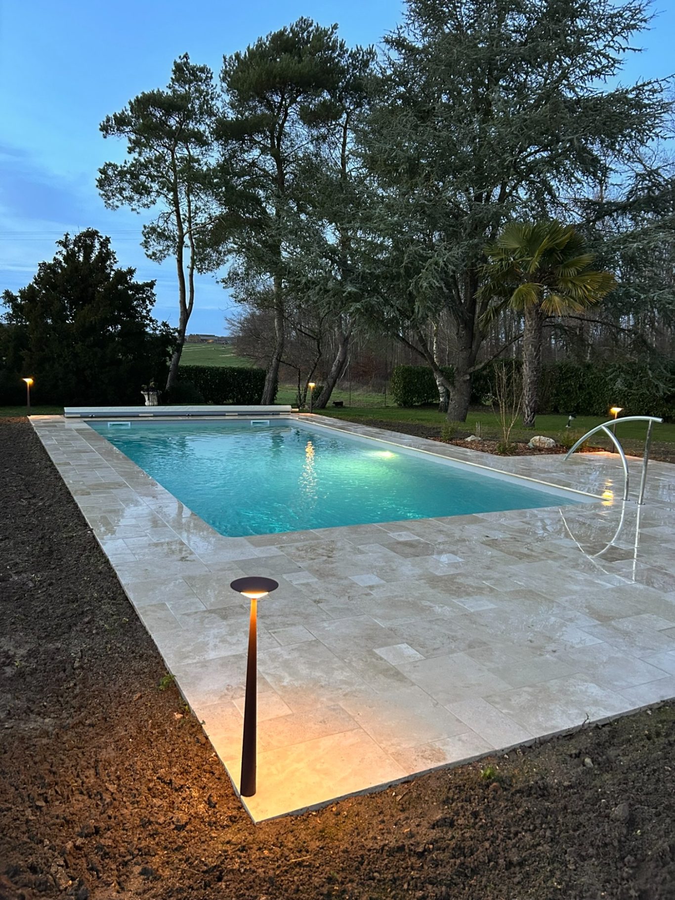 Bornes lumineuses pour terrasse Piscine entourée d’arbres, éclairée par des lampadaires, sous un ciel d’aube.