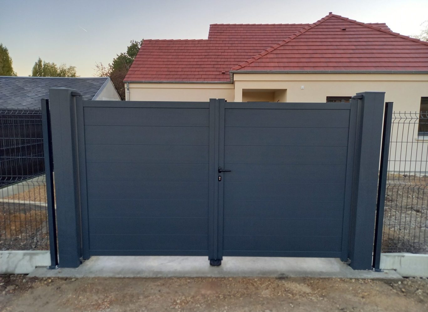 Panneau de clôture gris devant une maison avec toit rouge et terrain en béton.