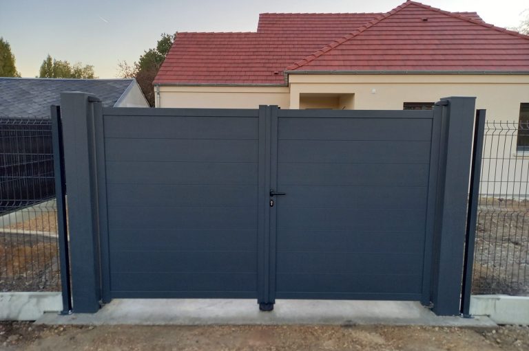 Portail en métal gris devant une maison avec un toit rouge. Terrassement en cours.