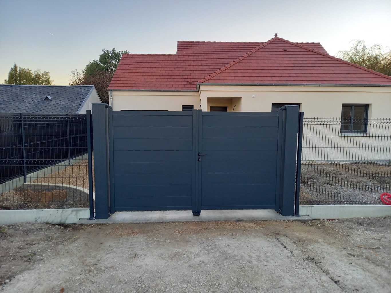 Pose des ventaux du portail Portail en métal gris devant une maison avec toit rouge, entourée de terrain en terre.