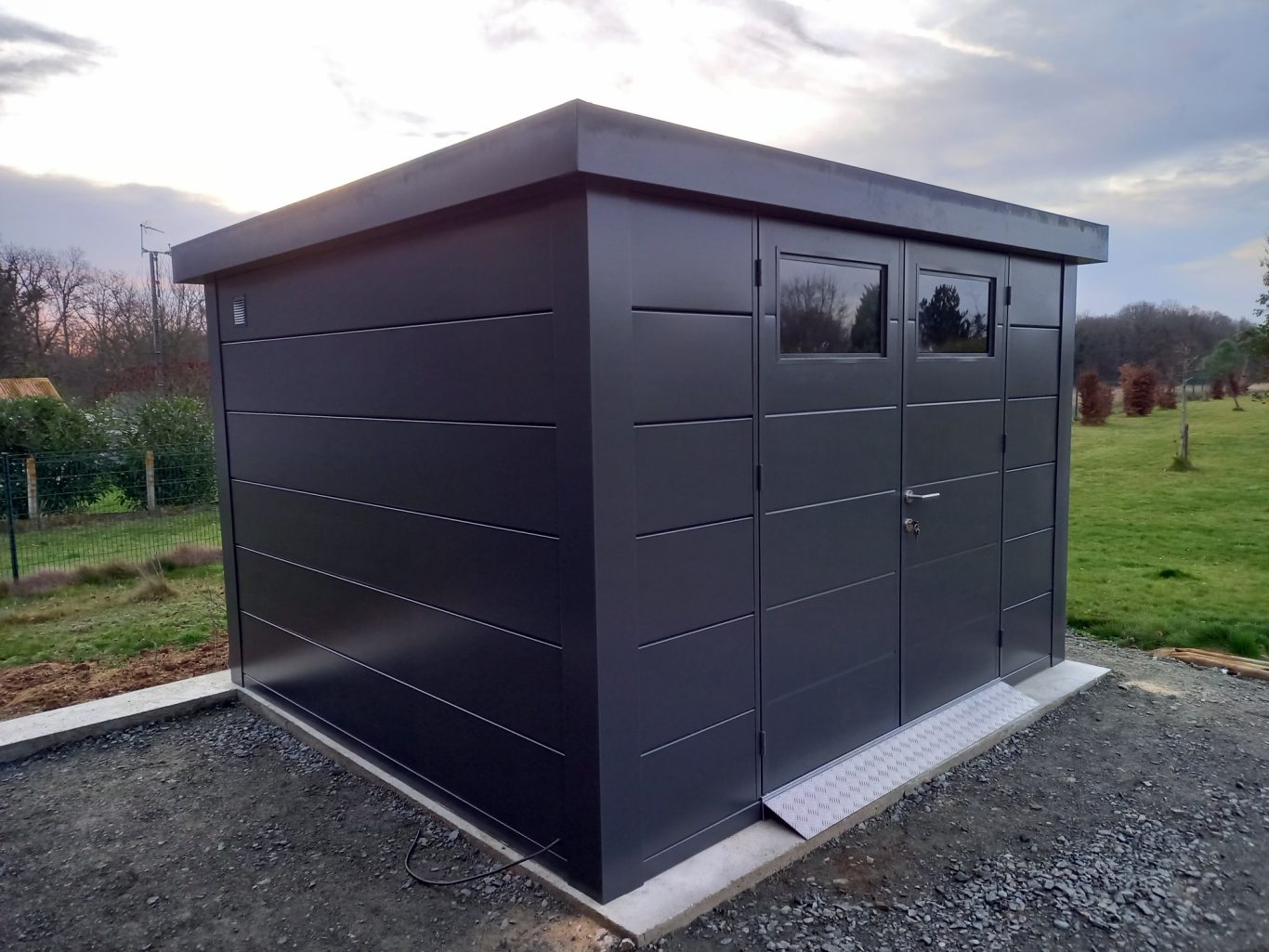 Abri de jardin isolé Abri de jardin moderne en métal, avec fenêtres et porte, sur un fond verdoyant.