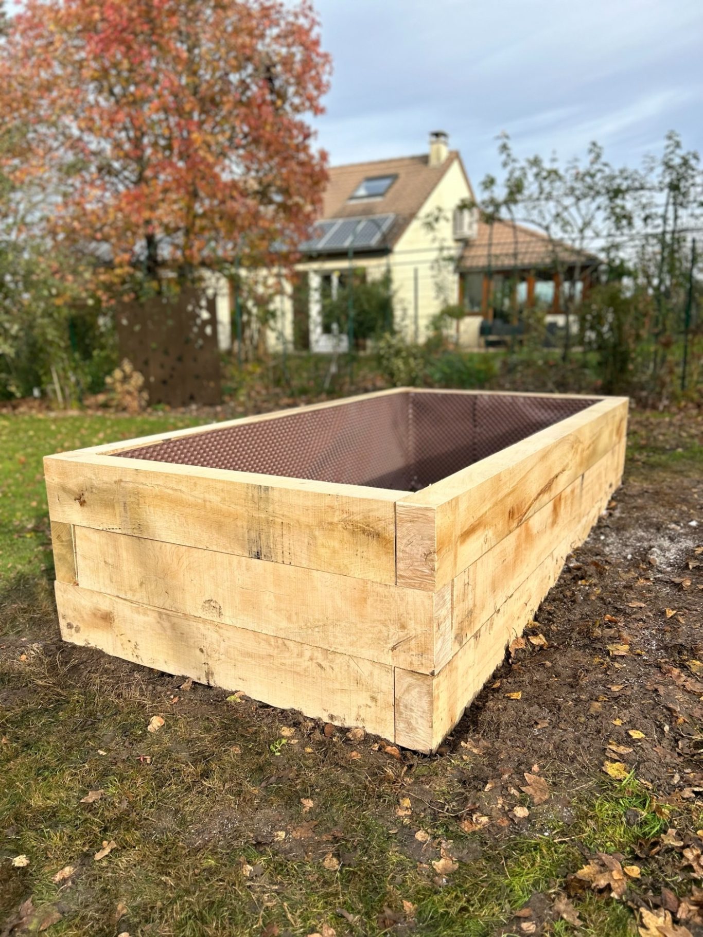 Bac pour carré potager Bac en bois non rempli, situé dans un jardin avec une maison en arrière-plan.