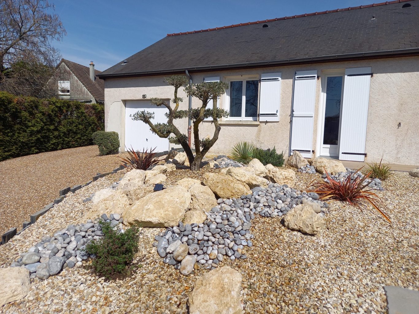 Rocaille Aménagement paysager avec des rochers, plantes et gravier devant une maison.