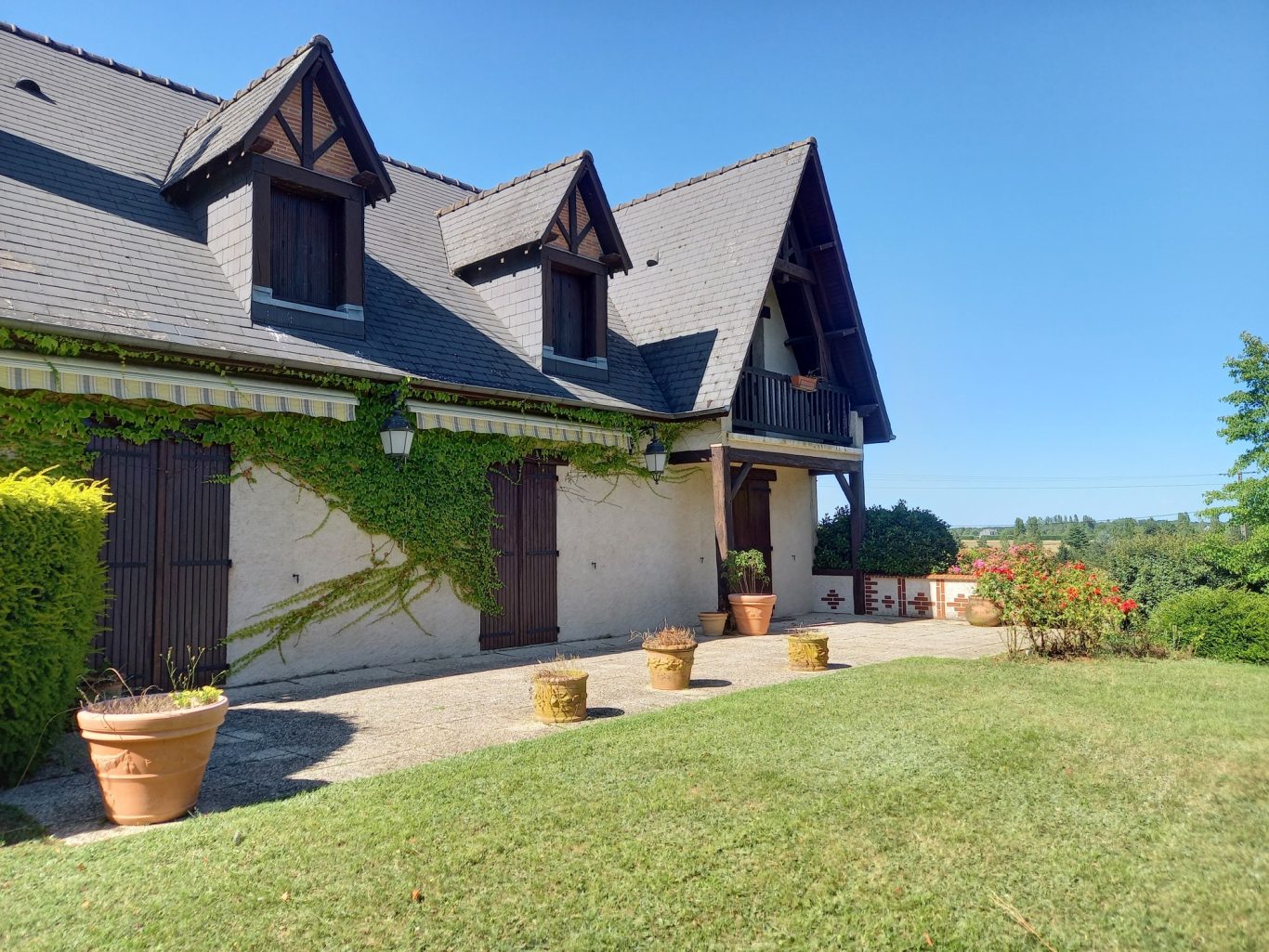 Tonte de pelouse Maison avec jardin, façade en pierre, arbres et pots de fleurs dans un cadre ensoleillé.