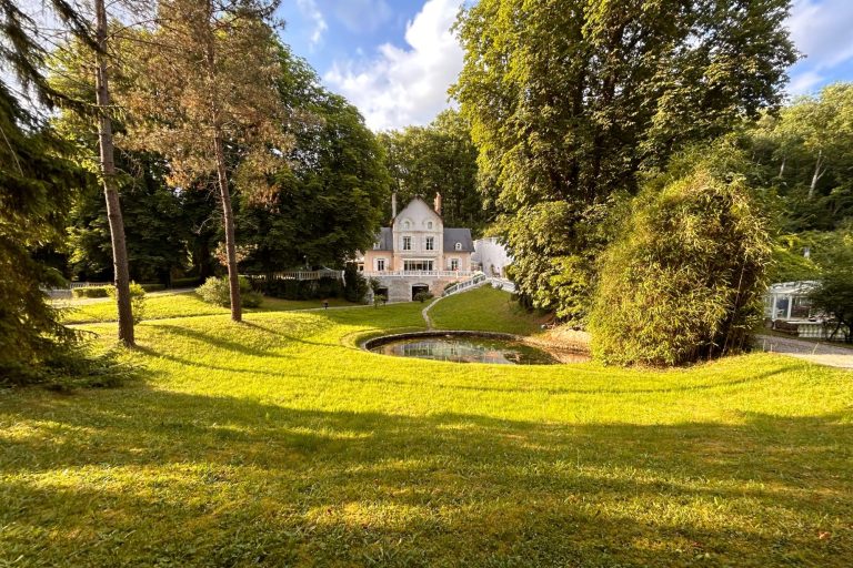 Maison blanche entourée d'arbres et de verdure, avec un étang au premier plan.