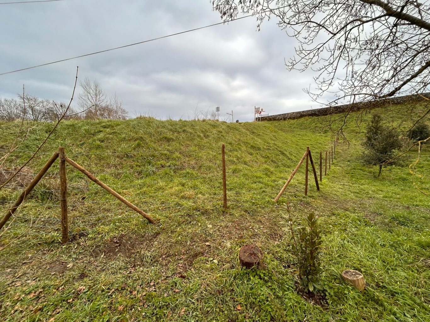 Clôture champêtre - Zone inondable Terrain herbeux avec des poteaux métalliques et une pente douce, sous un ciel nuageux.