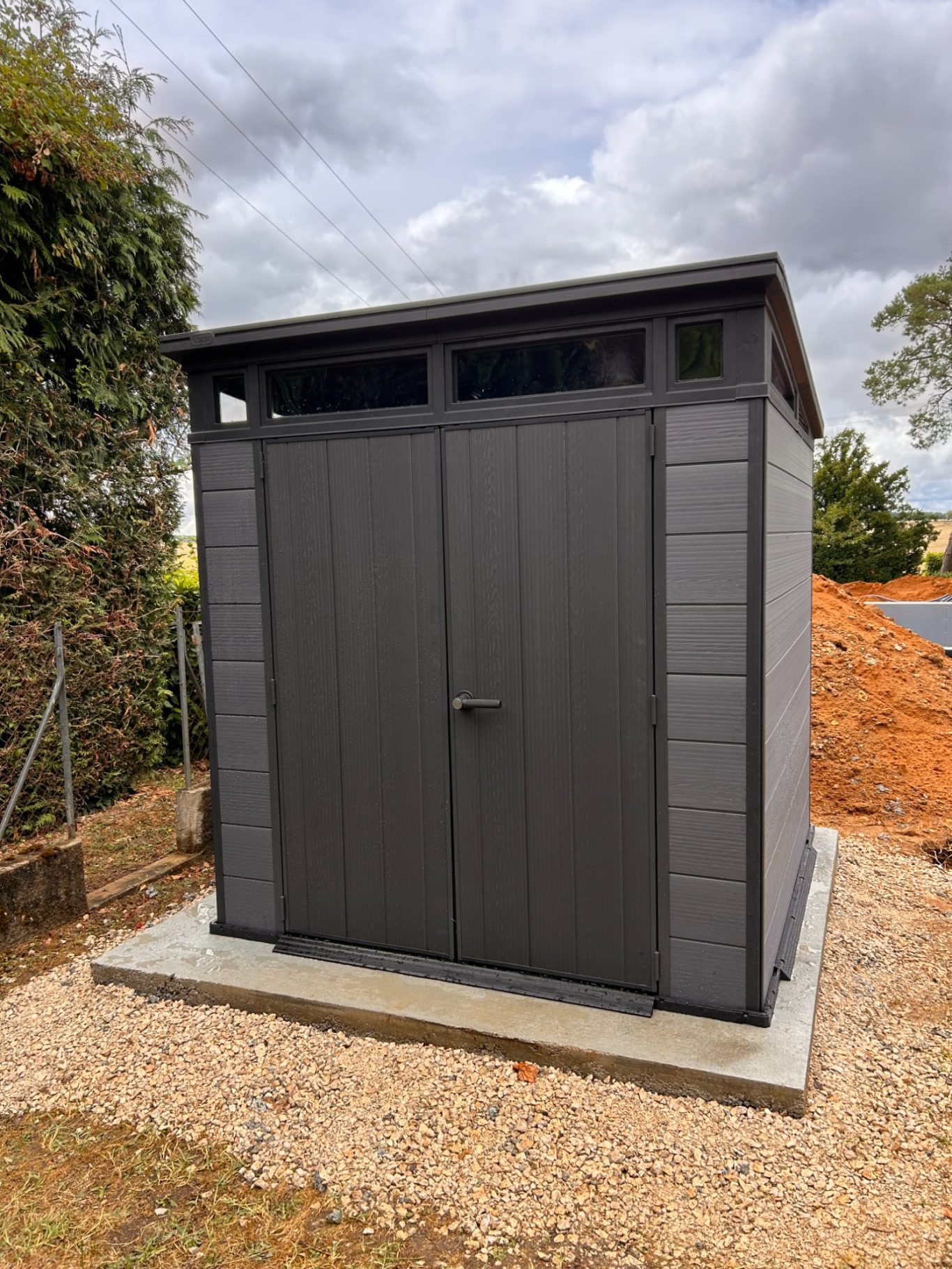 Abris de jardin en composite Cabane de jardin en métal gris, avec porte et fenêtres, sur une base en béton.
