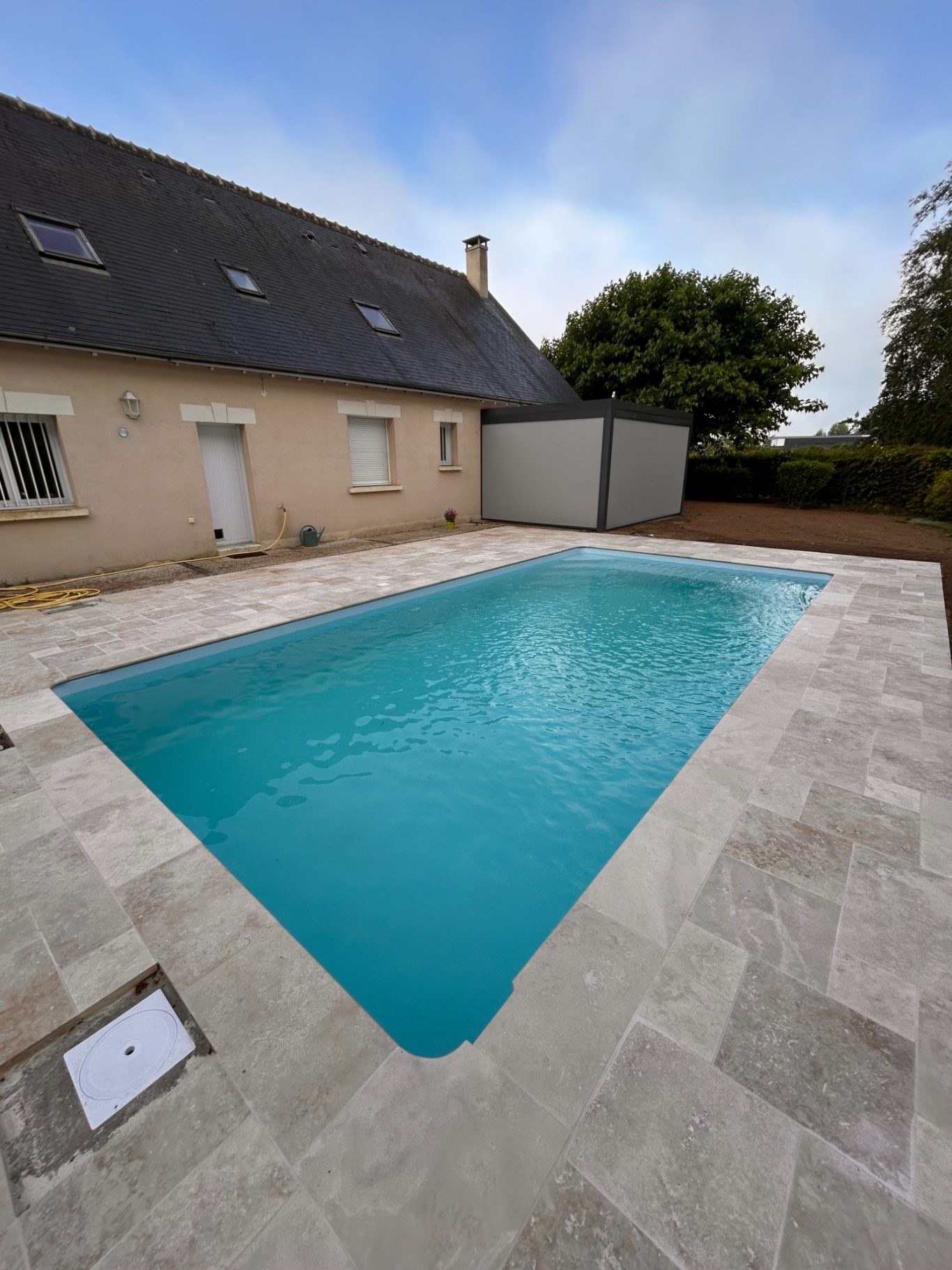 Travertin en Opus 4 formats Piscine turquoise entourée de dalles beige, près d'une maison à deux étages.